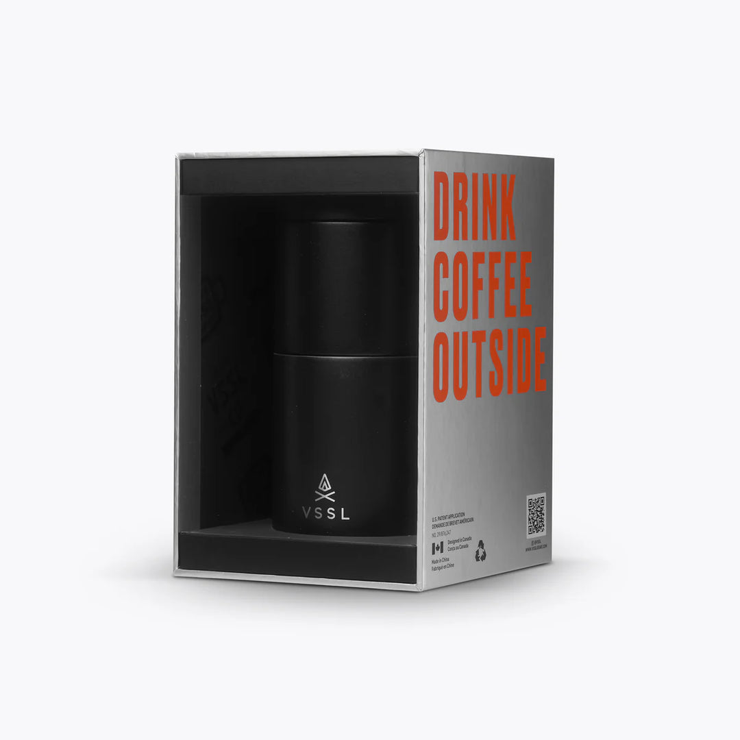 A black insulated coffee cup by VSSL, presented inside its open packaging box. The box is gray with bold orange text reading &