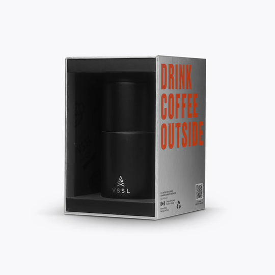 A black insulated coffee cup by VSSL, presented inside its open packaging box. The box is gray with bold orange text reading &