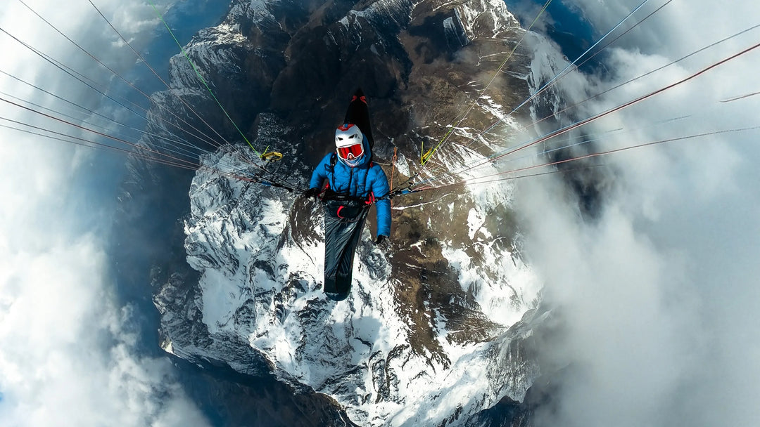 Person paragliding high above a mountainous landscape with clouds.