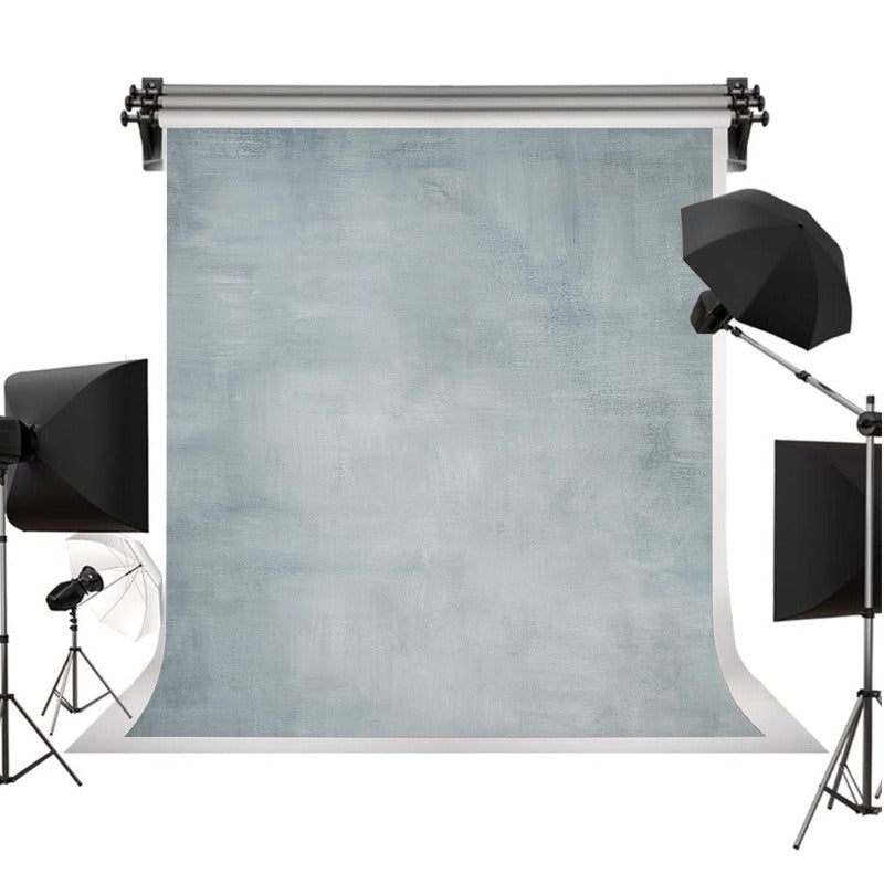 A photography backdrop set featuring a large textured light blue-gray backdrop roll suspended on a stand, surrounded by studio lighting equipment including softboxes and an umbrella on a stand against a white background.