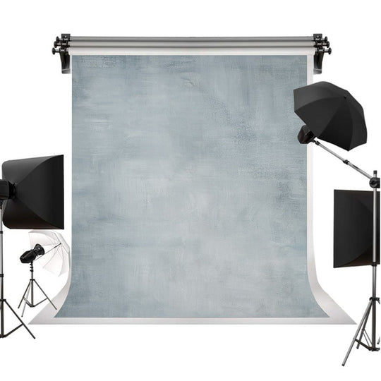 A photography backdrop set featuring a large textured light blue-gray backdrop roll suspended on a stand, surrounded by studio lighting equipment including softboxes and an umbrella on a stand against a white background.