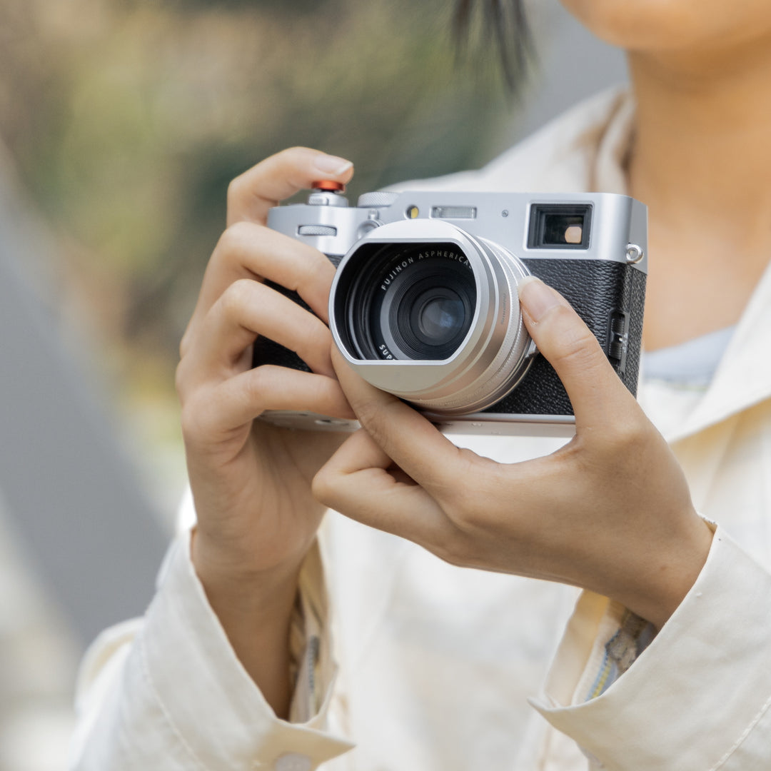 Photographer holding a silver FUJIFILM X100 series camera fitted with SmallRig silver lens hood and UV filter.