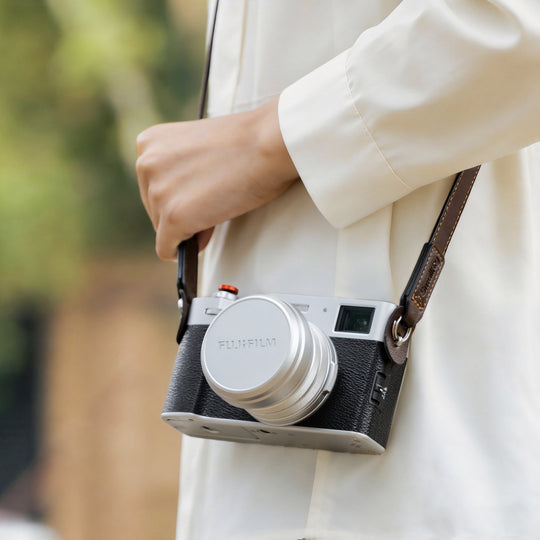 Silver FUJIFILM X100 series camera with original lens cap and SmallRig leather shoulder strap carried over the shoulder.