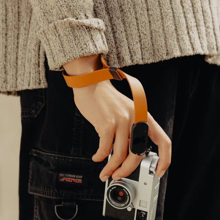 Person holding a vintage camera with an orange strap, wearing a textured sweater and dark pants.
