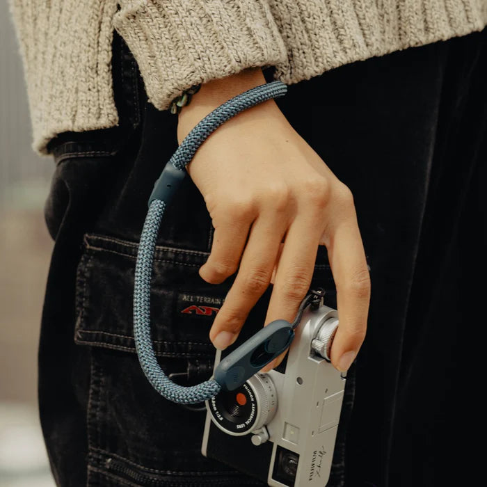 Person holding a vintage camera with a blue strap, wearing a beige sweater and dark pants.
