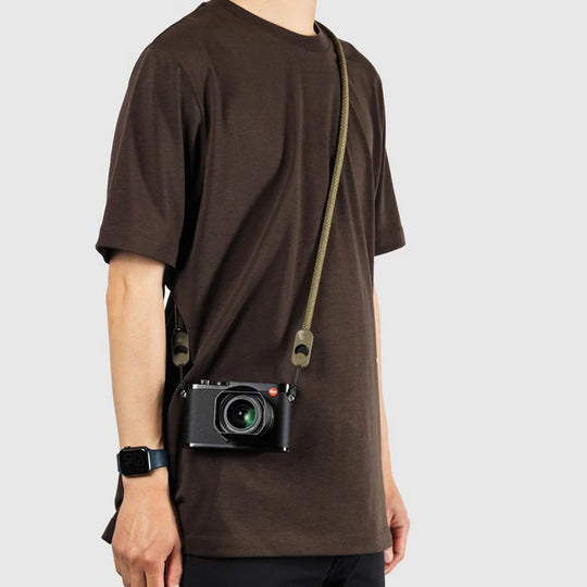 Person wearing a brown t-shirt with a camera attached to a strap over their shoulder on a light gray background